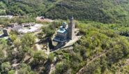 Aerial view of Tsarevets Fortress in Veliko Tarnovo, Bulgaria, showing the stone cathedral with a blue dome and bell tower, surrounded by greenery and ruins.