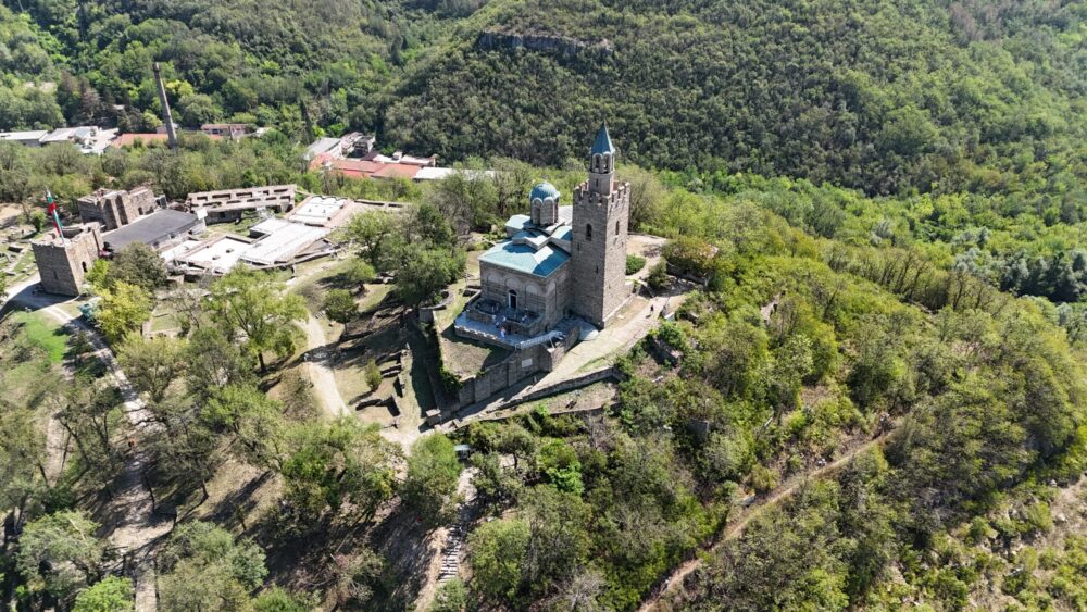 Aerial view of Tsarevets Fortress in Veliko Tarnovo, Bulgaria, showing the stone cathedral with a blue dome and bell tower, surrounded by greenery and ruins.